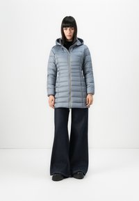 Vrouw in een lichtblauwe gewatteerde jas met capuchon, zwarte wijde broek en zwarte chunky schoenen, staande tegen een witte achtergrond.