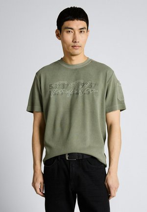 Mężczyzna w oliwkowo-zielonym T-shircie z krótkim rękawem z napisem z przodu i czarnych spodniach stoi na jednolitym tle.