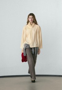 Vrouw draagt crèmekleurige asymmetrische top met knopen en bruine broek, draagt een rood gebreid item en loopt op een effen vloer binnenshuis met een grijze muur op de achtergrond.