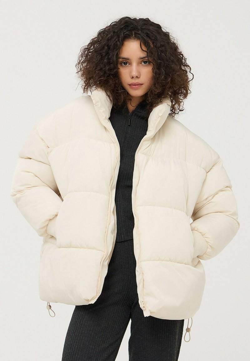 Giacca puffer color crema con design a righe orizzontali, colletto alto, chiusura con zip e polsini elasticizzati. Tessuto morbido e liscio.