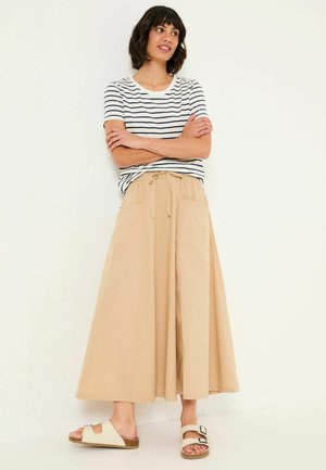 Woman with short dark hair standing against white wall, wearing white and black striped t-shirt, beige wide-leg pants, and cream sandals.