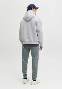 Grauer Kapuzenpullover mit großer Kapuze, kombiniert mit hellgrauen Slim-Fit-Sweatpants mit seitlichen Streifen. Das Model trägt weiße Sneaker und eine marineblaue Mütze.