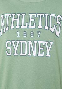 Maglietta di cotone verde con testo bianco e azzurro chiaro che recita "ATHLETICS SYDNEY 1987." Semplice scollo a girocollo e vestibilità comoda.