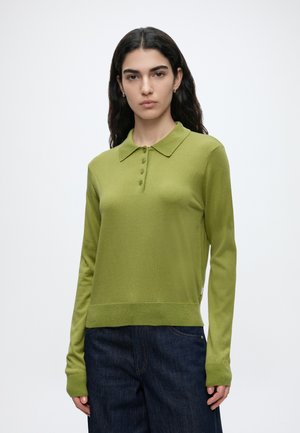 Femme aux longs cheveux foncés portant un polo manches longues vert clair et un jean bleu foncé, debout devant un fond blanc uni.