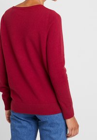 Roter, gestrickter Pullover mit rundem Ausschnitt und gerippten Bündchen. Getragen zu blauen Jeans, die eine lockere Passform und weiche Textur zeigen.