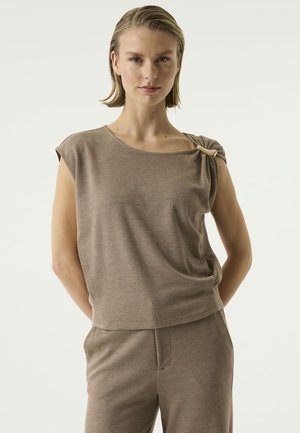Frau mit kurzen Haaren, die ein ärmelloses Top in Taupe mit einer dekorativen Schnalle an der Schulter und passende Hosen trägt, steht mit den Händen hinter dem Rücken.
