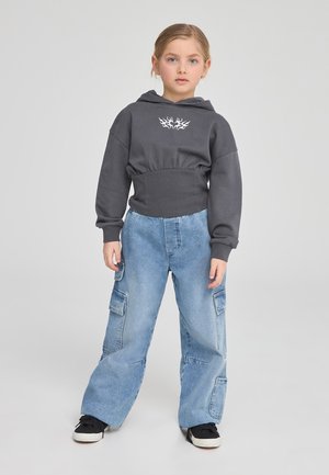 Felpa grigia corta con cappuccio e cordoncino, caratterizzata da un design di fiamme bianche; abbinata a pantaloni cargo larghi di colore blu chiaro con tasche laterali.