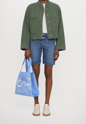 Osoba w zielonej krótkiej kurtce, białej bluzce, dżinsowych szortach, beżowych sneakersach, trzymająca niebieską torbę na zakupy z kolorowymi ilustracjami związanymi z plażą.