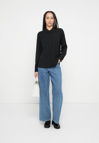 Black long-sleeve shirt with a collar, paired with wide-leg blue denim jeans. White handbag, black shoes, model stands against a plain background.