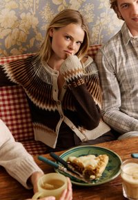 Gilet en tricot avec des motifs en zigzag marron, beige et orange, associé à un haut marron uni. Assiette verte avec une tarte et des ustensiles sur une table en bois.