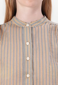 Camisa riscada em bege e preto, com colarinho tipo banda e riscas verticais, apresentando quatro botões na frente e uma textura suave.