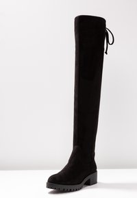 Bota negra por encima de la rodilla hecha de material de ante, con tacón cuadrado, punta redondeada y detalle de lazo ajustable en la parte superior.