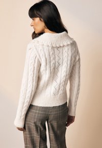 Beli pulover s pletenimi kabelskimi vzorci in velikim zaobljenim ovratnikom. Ima teksturirane vzorce in rob z rebrasto strukturo. Oblečen s karirastimi hlačami.