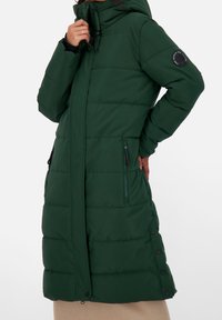 Abrigo acolchado verde con capucha, con bolsillos laterales con cremallera, diseño con paneles y botones de presión a lo largo del lateral. Detallado con un parche de logo.