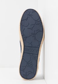 Semelle en caoutchouc bleu marine avec motif floral ; couche de type jute autour du bord. Présente le logo "ROXY" au centre.