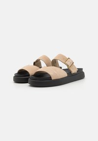 Beige suede sandals with two wide straps and a gold buckle detail, resting on a black flat sole. Textured upper for comfort.