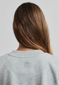 Sudadera gris con cuello redondo y logo bordado en la parte trasera. La tela parece suave con una textura lisa.
