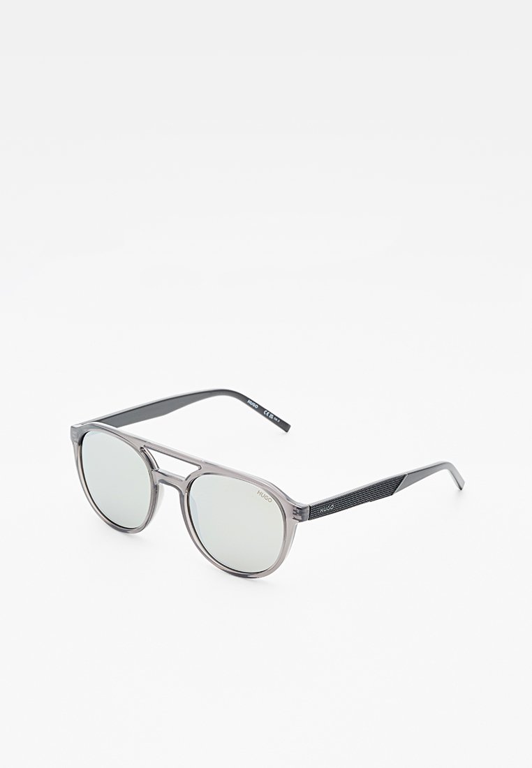 Runde graue Sonnenbrille mit dünnen, durchscheinenden Rahmen und schwarzen strukturierten Bügeln, mit der Markenbezeichnung "HUGO" auf dem Glas und den Schläfen.