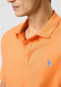 Orange polo shirt with a textured fabric, featuring a blue embroidered logo on the left chest and a contrasting collar.