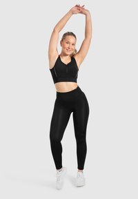 Reggiseno sportivo nero con zip frontale, abbinato a leggings neri a vita alta. Entrambi hanno una texture liscia e pochi accenti bianchi.