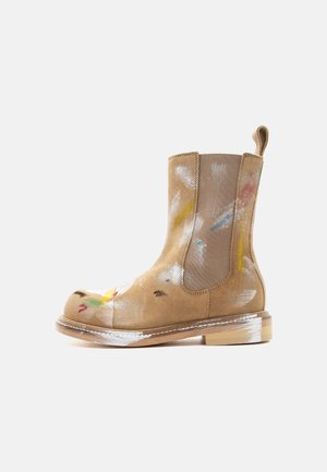 Bottine Chelsea en daim camel avec panneaux élastiques sur les côtés, languette de tirage et éclaboussures de peinture multicolores sur la tige et la semelle.