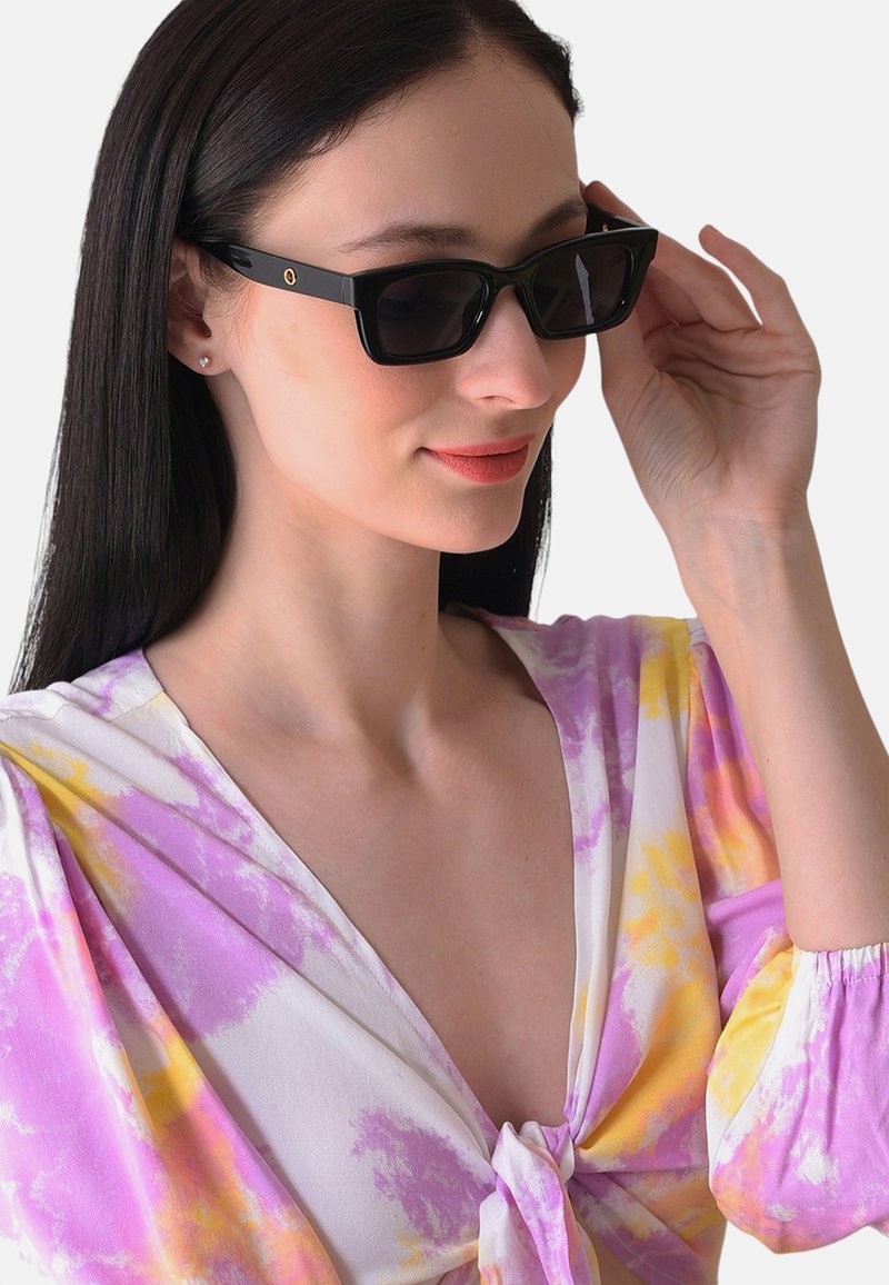 Zwarte rechthoekige zonnebril met donkere lenzen, voorzien van een kleine gouden accent aan de zijkant, gedragen met een kleurrijke tie-dye blouse.