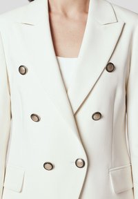 Dwurzędowa biała marynarka z wyłogami, wykonana z gładkiego materiału, z pięcioma okrągłymi złotymi guzikami i kieszeniami z przodu.