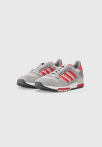 Grijze suède sneakers met rode accenten, voorzien van drie strepen, witte tussenzool en zwarte rubberen buitenzool. Vetersluiting met gewatteerde kraag.