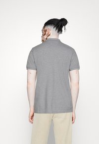 Osoba s pletenými vlasy svázanými do drdolu, oblečená v šedém tričku s krátkým rukávem a světlých kalhotách, viděná zezadu na bílém pozadí.