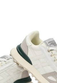 Zapatillas blancas con acentos verdes, suelas texturizadas y detalles de ante gris claro. Incluye una etiqueta en la lengua con información del producto.