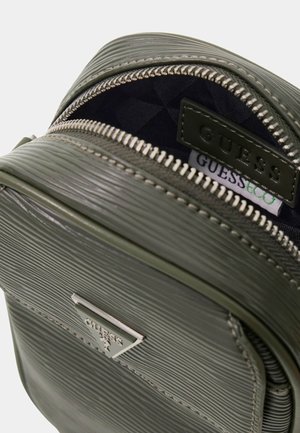 Zielony teksturowany skórzany mini plecak Guess z częściowo otwartym srebrnym zamkiem błyskawicznym, pokazującym czarną wyściółkę i logo marki wewnątrz.