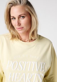 Gele sweatshirt met wit reliëftekst waarop "POSITIVE HEARTS" en "RUE DE PARIS" staat. Zachte stof, ronde halslijn en losse pasvorm.
