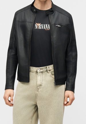 Man wearing unzipped black leather jacket over black Nirvana t-shirt and light beige jeans, standing with hands relaxed by sides.