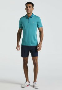Chemise polo turquoise avec un motif texturé, trois boutons et un col contrastant ; assortie avec un short marine et des baskets blanches.