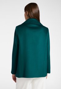 Donna con capelli castani lisci che indossa un cappotto di lana turchese e pantaloni bianchi, voltata di spalle su uno sfondo chiaro e neutro.