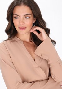 Blusa beige chiaro con scollo a V, maniche lunghe con dettagli a bottoni. Tessuto morbido, texture liscia e vestibilità rilassata.