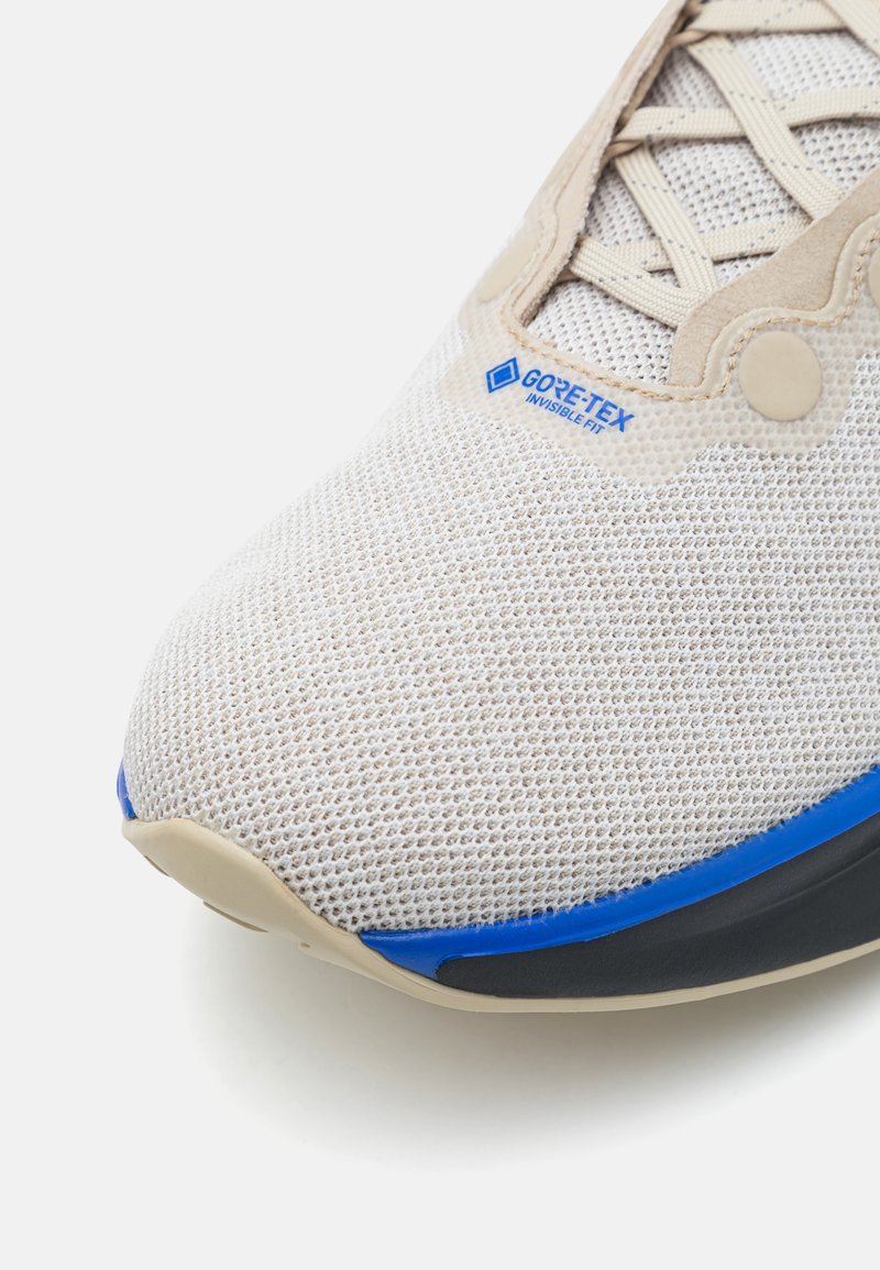 Zapatilla ligera con parte superior de malla beige, acentos azules y suela negra. Presenta la marca Gore-Tex en el costado.