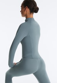 Femme avec les cheveux en chignon portant une tenue de sport ajustée bleu clair, vue de profil, dans une posture d'étirement avec un genou plié et les mains reposant sur la cuisse.