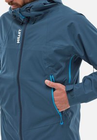 Veste imperméable bleue avec fermeture éclair, capuche réglable et accents bleus contrastés. Comprend des poches latérales et un design épuré.