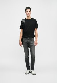 Νεαρός άνδρας με μαύρη μπλούζα, γκρίζα τζιν, λευκά αθλητικά παπούτσια, κρατώντας μια μαύρη τσάντα ώμου, με φόντο λευκούς τοίχους.