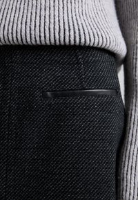 Musta teksturoidut housut, joissa on diagonaalinen kudonta, kapea istuvuus ja kapea musta nahkainen korostetasku takana.