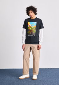 T-shirt preta com uma estampa floral colorida e a frase "Move com a Natureza", sobreposta a uma camisa de manga longa branca; combinada com calças bege e sapatilhas brancas.