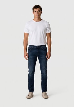 Mann, der frontal steht, trägt ein schlichtes weißes T-Shirt, dunkelblaue Jeans und beige Sneakers vor einem hellgrauen Hintergrund.
