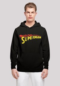 Fekete kapucnis pulóver, amelyen egy sárga és piros "Superman" logó található a mellrészen. Kenguru zsebe van és állítható húzózsinórral ellátott kapucnija.