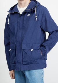 Giacca a vento blu navy realizzata in tessuto leggero e testurizzato. Presenta un cappuccio, cordini regolabili e due tasche frontali.