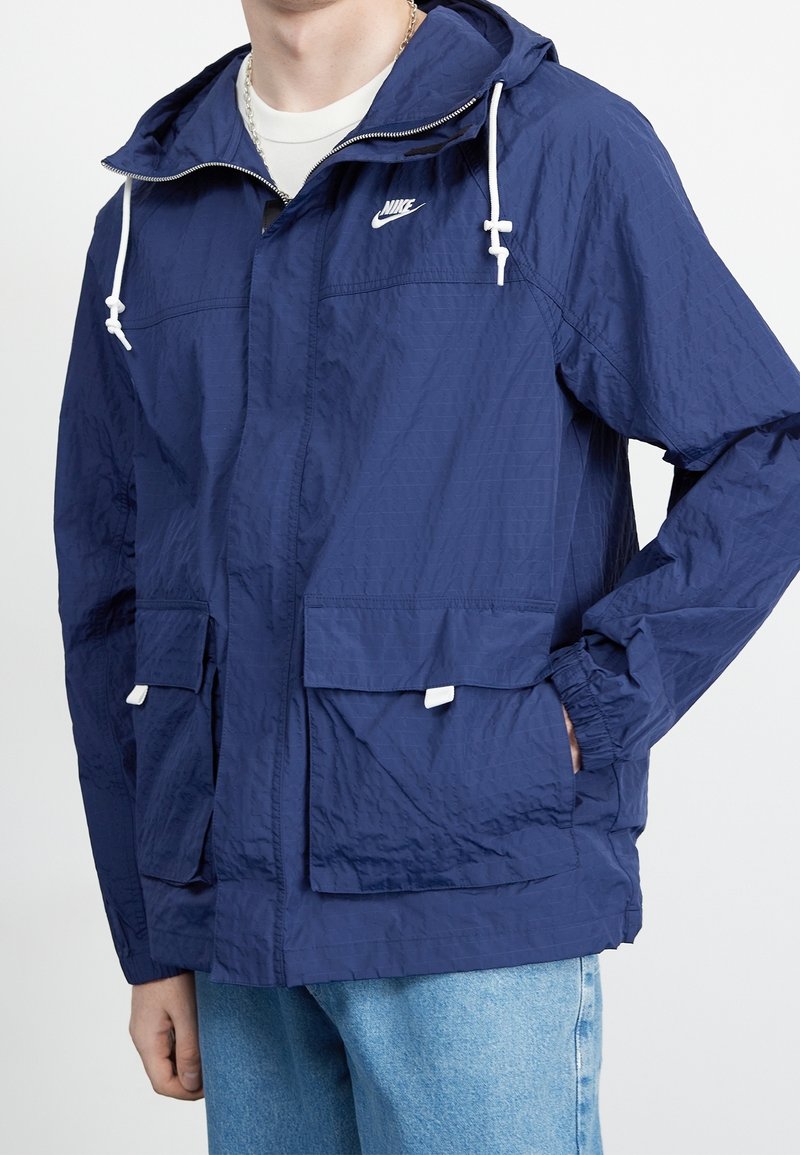 Giacca a vento blu navy realizzata in tessuto leggero e testurizzato. Presenta un cappuccio, cordini regolabili e due tasche frontali.