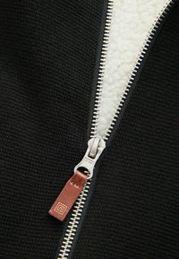 Must mustriga jakk musta värviga, millel on hõbedane tõmblukk ja nahast tõmbluku kinnituse osa, paljastades valge fliisvoodri. Tekkiv tekstuurne kude on nähtav.