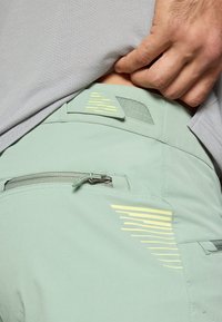 Svijetlozelene hlače s patentnim zatvaračem, s žutim naglascima na pojasu i glatkom, laganom tkaninom.