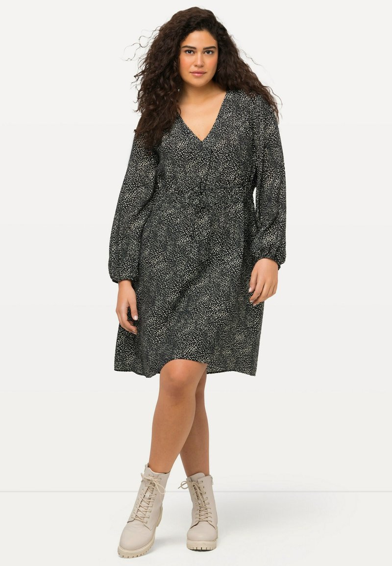 Vestido negro con manchas blancas, escote en V, mangas largas abullonadas, cintura ajustada, hasta la rodilla, combinado con botas beige con cordones.