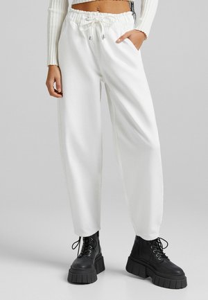 Pantalons de survêtement blancs, amples, avec une taille élastique et un cordon de serrage. Comprend des poches latérales et des ourlets côtelés. Associés à des bottes noires imposantes.